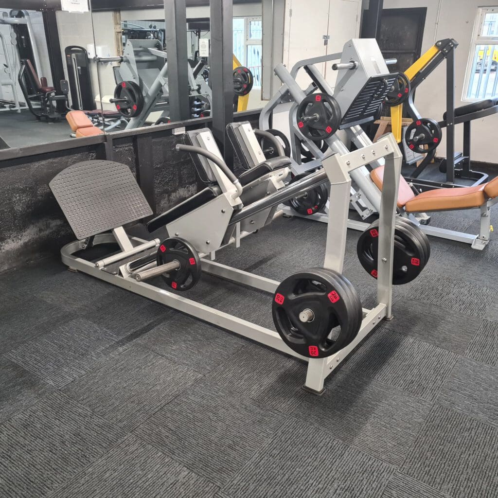 Leg press machine in a gym with several weight plates on it. The machine is positioned on a carpeted floor near a mirror and other fitness equipment, including a seated bench and weight stack machine in the background.
