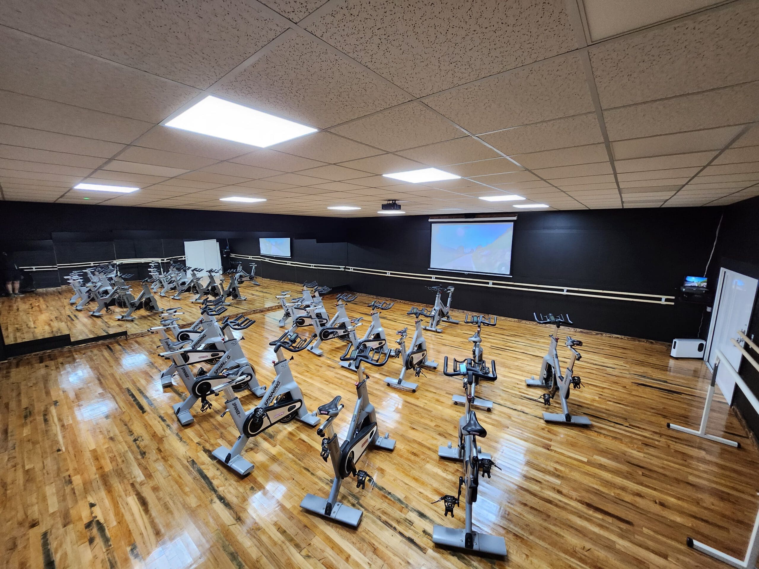 A spacious indoor cycling studio with multiple stationary bikes arranged in rows on a polished wooden floor. The room has a mirrored wall, a large screen at the front, and soft overhead lighting.