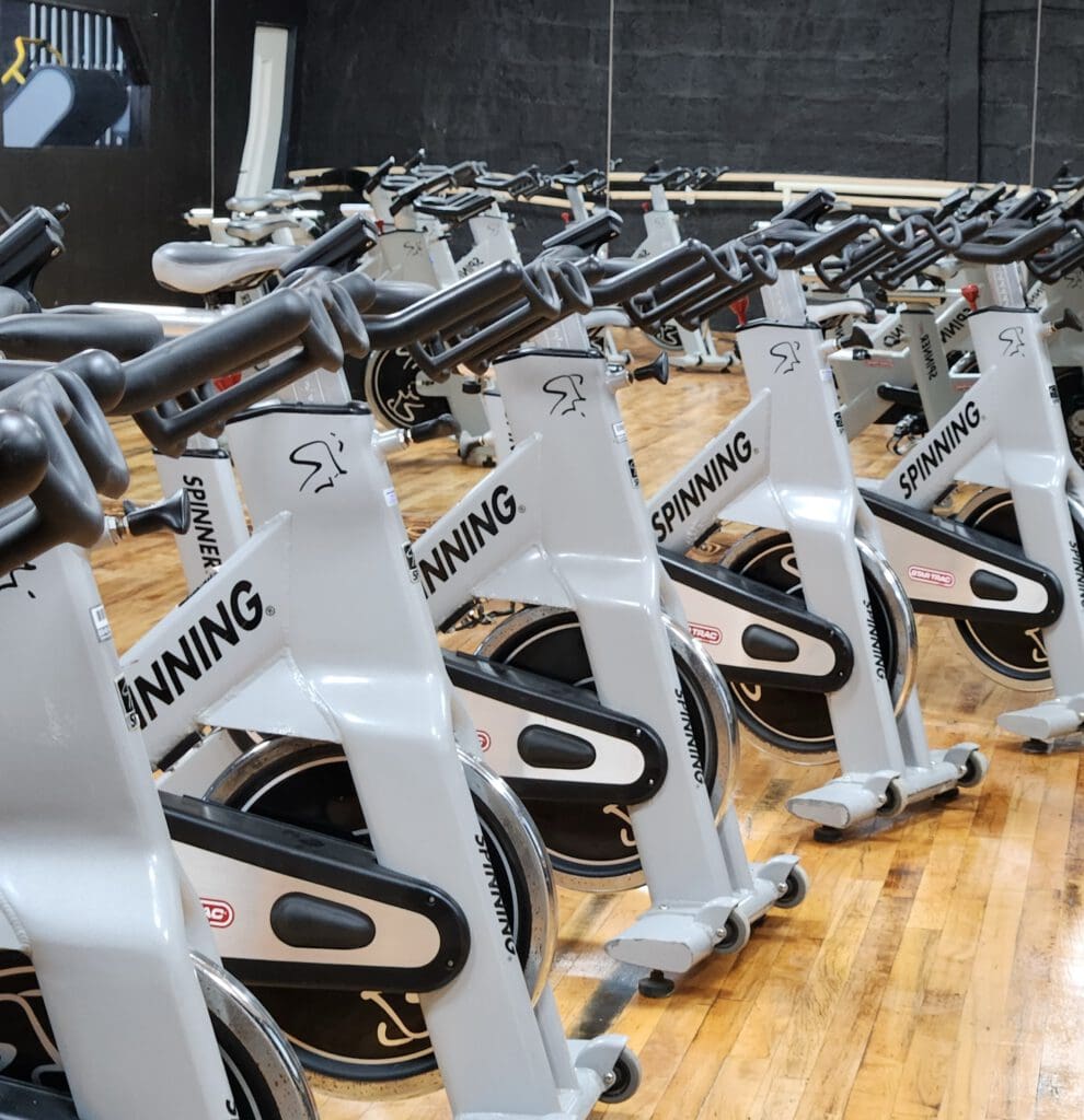 A row of stationary spinning bikes in a gym with wooden floors. The bikes are silver with black handles and are aligned in straight formation. A large mirror is visible in the background, reflecting the room's interior.