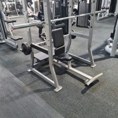 A gym setting with a bench press station featuring a padded bench, barbell rack, and barbells with weights on both ends. Other gym equipment is visible in the background on a carpeted floor.