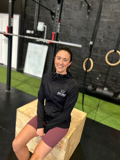 A person in a black hoodie and maroon shorts sits on a wooden box in a gym. Behind them are gymnastic rings and a pull-up bar, with a black brick wall in the background.