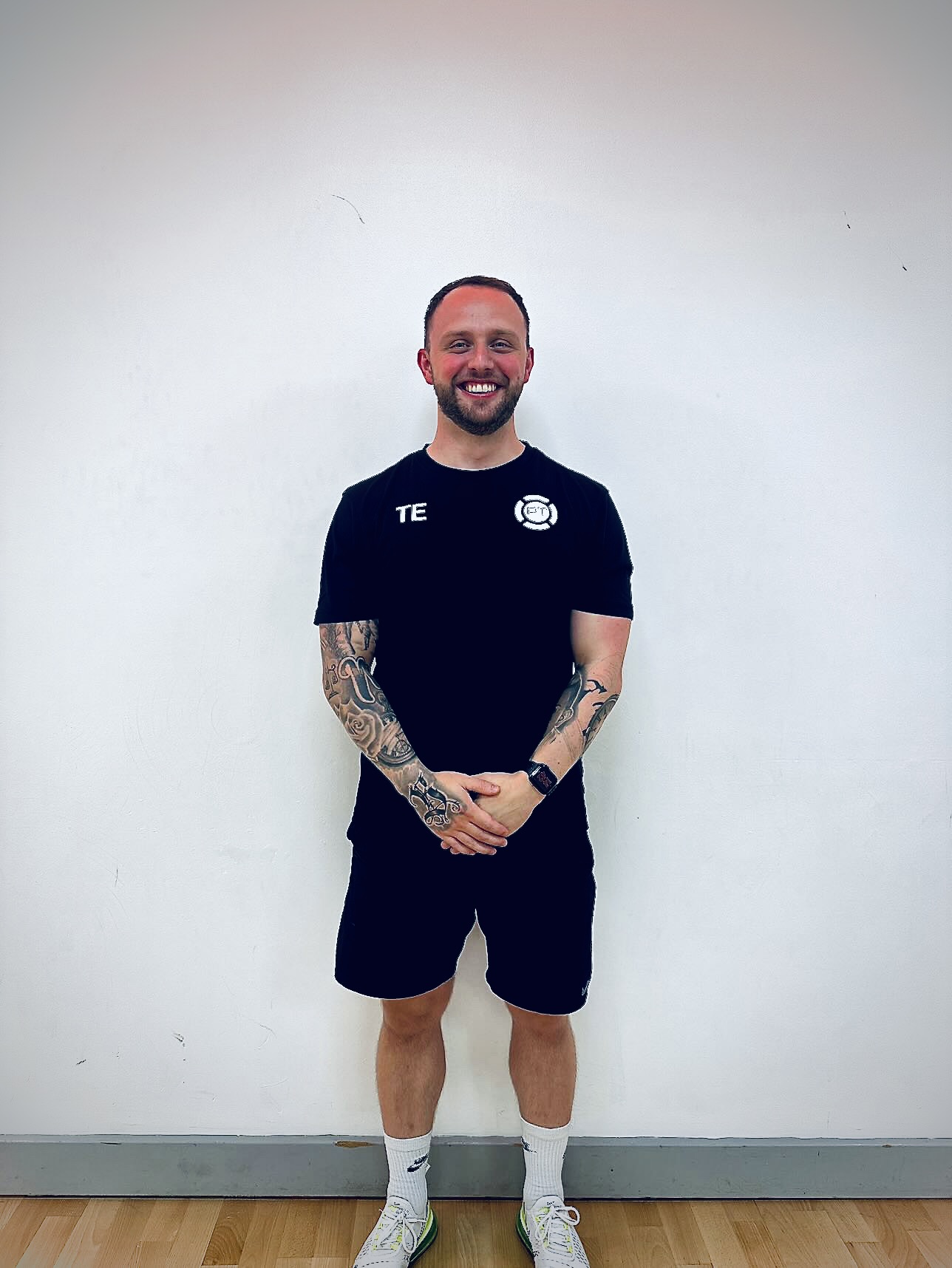 A smiling man with tattoos on both arms stands against a plain white wall. He is wearing a black t-shirt, black shorts, white socks, and white sneakers, with his hands clasped in front of him.