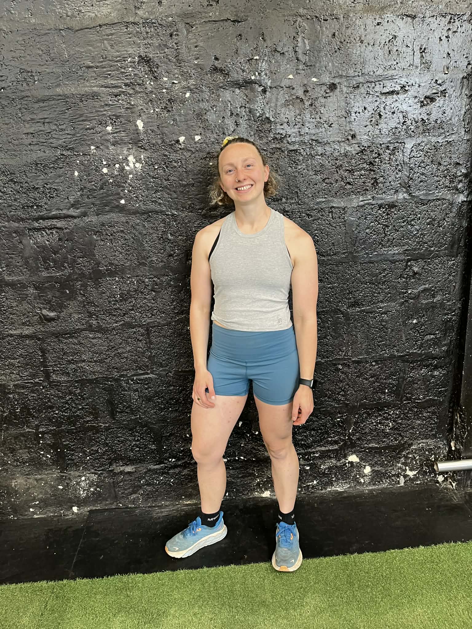 A woman in a gray sleeveless top and blue shorts stands smiling in front of a textured black wall on a gym floor with green turf. She is wearing blue sneakers and a wristwatch.