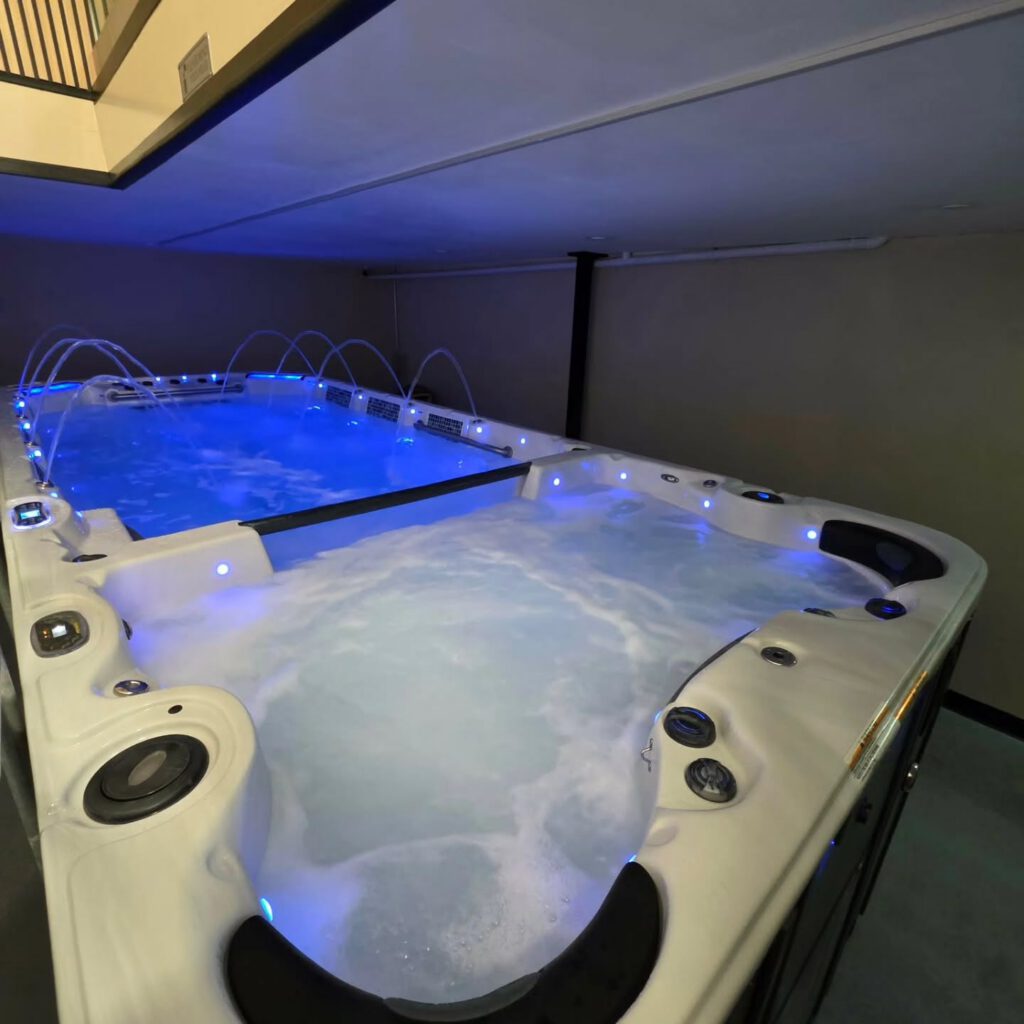 Large indoor hot tub with blue LED lights and water jets creating bubbles and streams, surrounded by beige walls and a low ceiling.