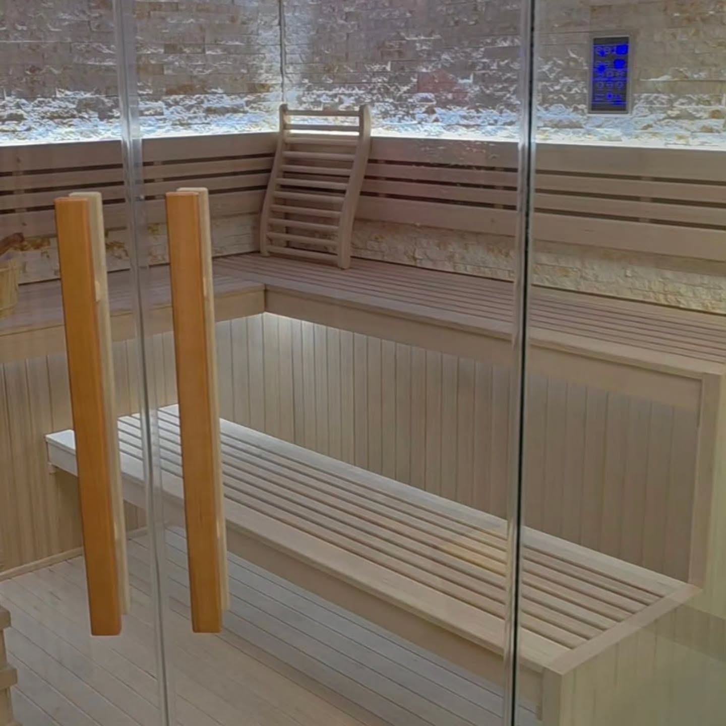 A modern wooden sauna with glass doors, a bench, a backrest on the upper level, stone walls, and a digital control panel on the wall.