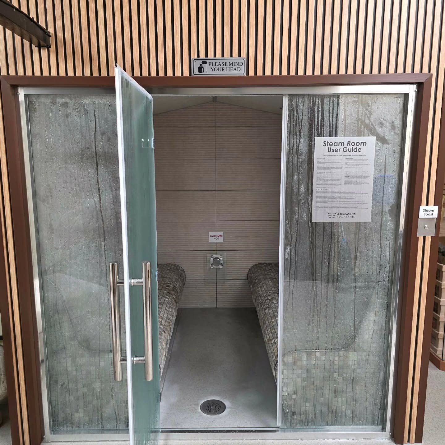 A steam room with glass doors slightly open, two tiled benches inside, a sign reading “PLEASE MIND YOUR HEAD” above, and a user guide posted on the right glass panel. Walls are wooden slats.