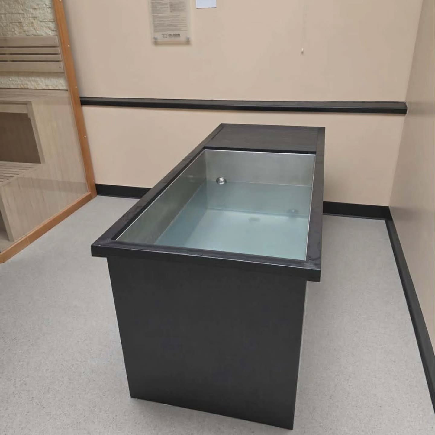 A rectangular black ice bath or cold plunge tub filled with water stands in a clean, beige room with light flooring and a wooden door to the left.