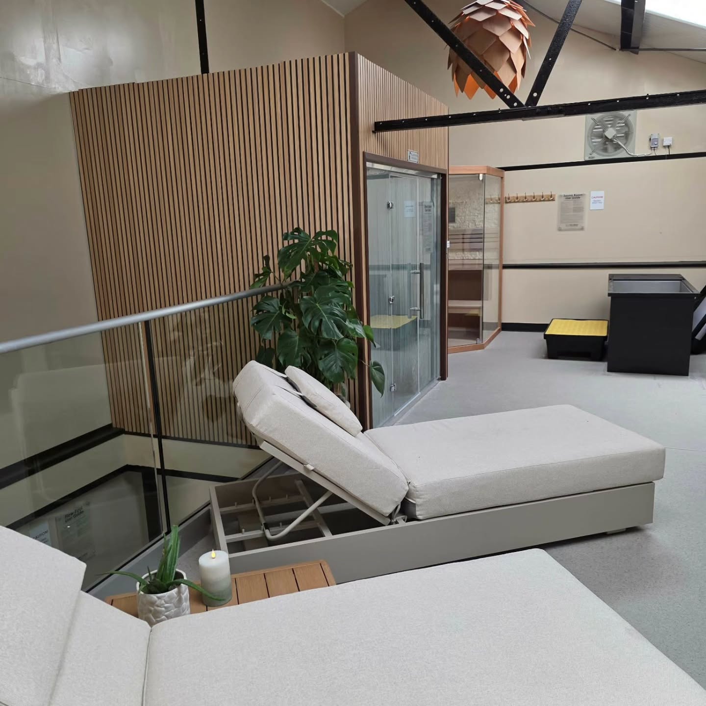 Two beige lounge chairs face a glass enclosure and wooden sauna in a modern, serene spa setting. A large green plant, small table, and candle add to the relaxing atmosphere. Black beams and soft lighting enhance the calm environment.