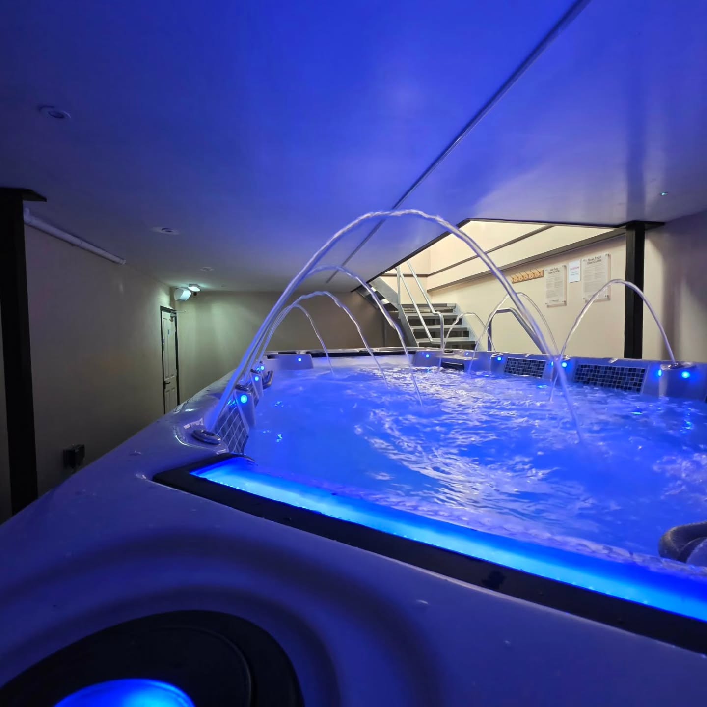 A close-up of a blue-lit indoor hot tub with water jets spraying arcs, set in a room with cream walls, ceiling lights, and a white door in the background.